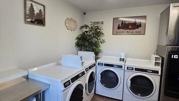 A laundry room with two front-loading washing machines, a dryer, and a refrigerator. There are wall decorations including a bear and a mountain scene.