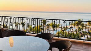 A balcony with a glass table and wicker chairs, overlooking a beach with palm trees and a clear sky. - Amazing 3-bedroom condo with pool, fitness room, in phenomenal Chileno Bay, Cabo (San José del Cabo)