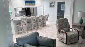 Uma sala de estar moderna com um sofá cinzento, uma poltrona estampada e uma área de jantar, com uma cozinha ao fundo.
