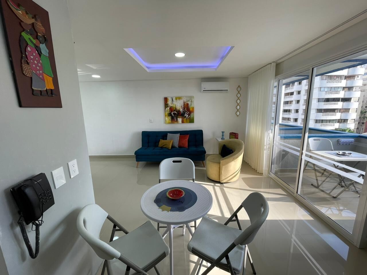 Un salon moderne avec un canapé bleu, une table ronde surmontée d’un saladier, des chaises blanches et une cloison en verre donnant sur un balcon.