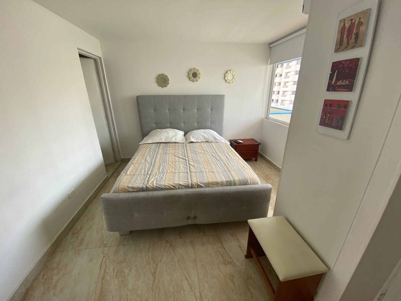 A bedroom with a large bed, a bedside table, a bench, and a window with artwork on the wall.
