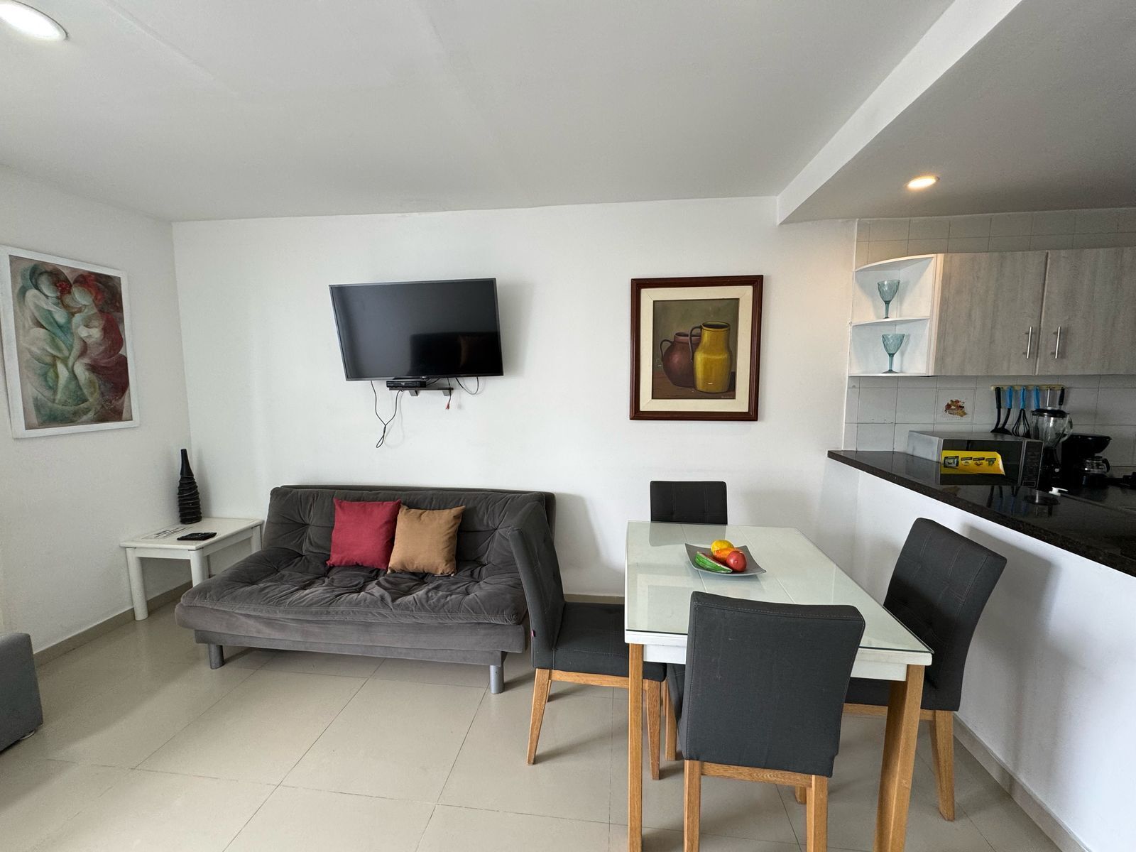 A modern living space with a grey sofa, a dining table with chairs, a flat-screen TV mounted on the wall, and a kitchen area with cabinets and appliances.