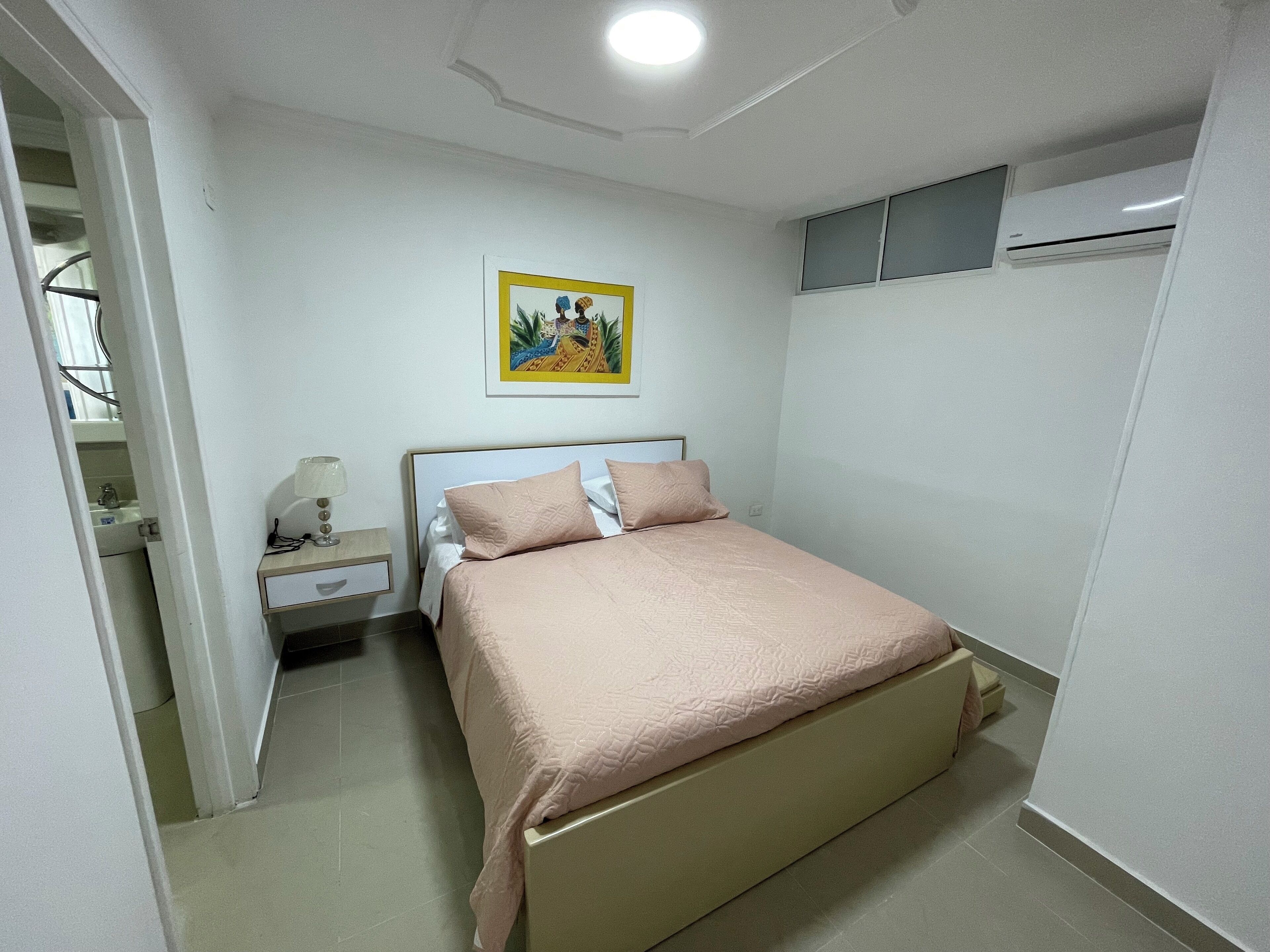 A bedroom with a bed, a nightstand, a wall-mounted air conditioner, and a painting.