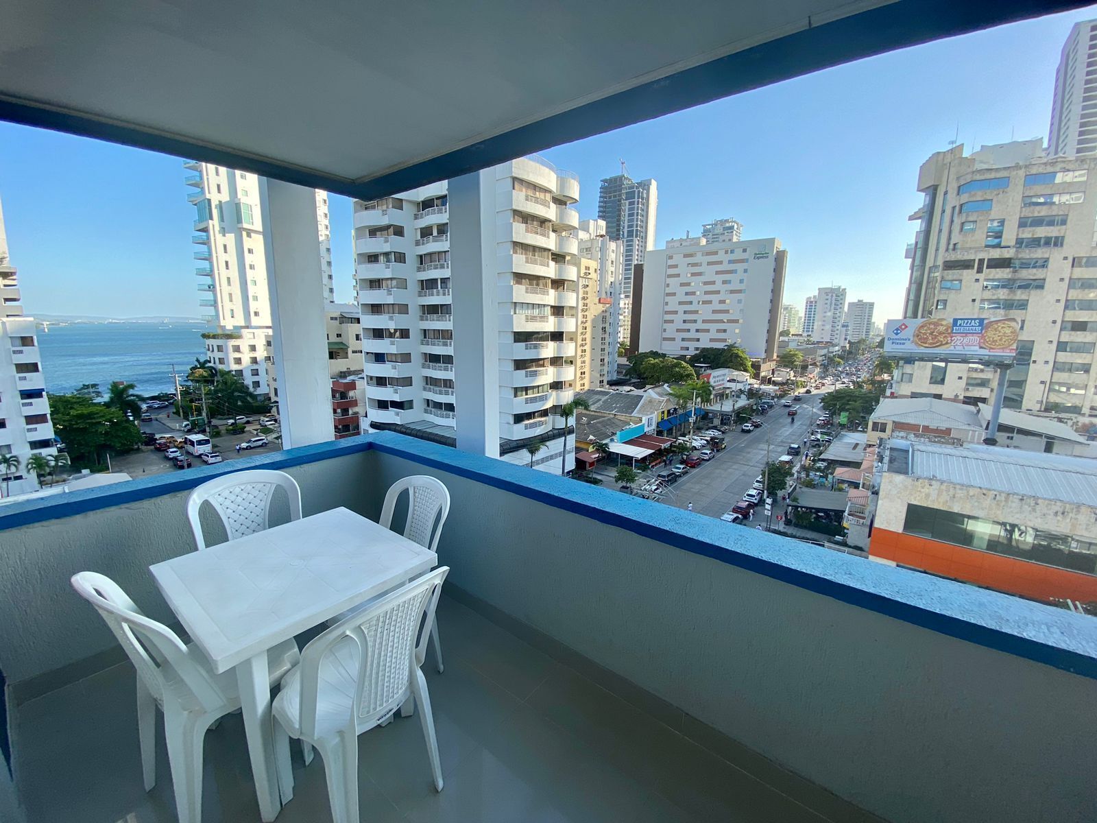 A balcony with a table and chairs, overlooking a cityscape with tall buildings and a view of the sea.