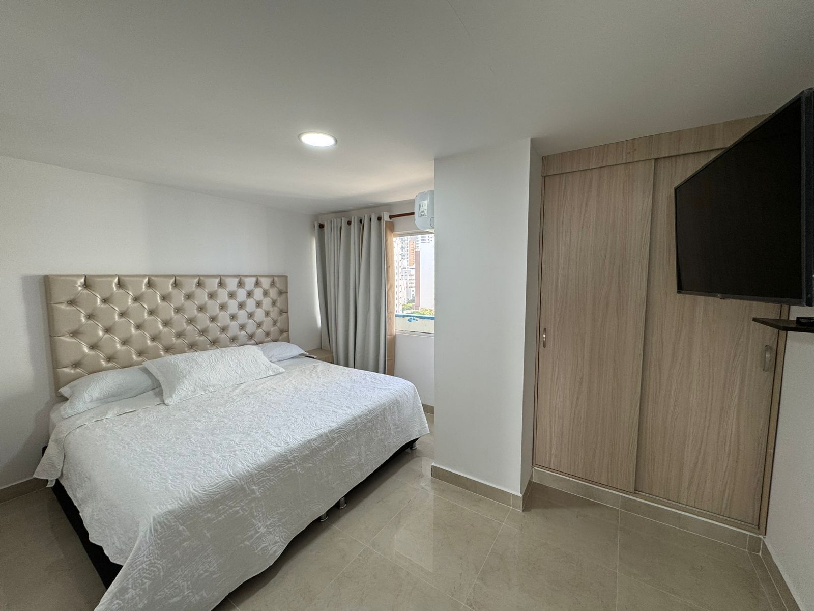 Una camera da letto con testiera trapuntata, letto con lenzuola bianche, televisore a schermo piatto montato a parete e una porta scorrevole in legno.