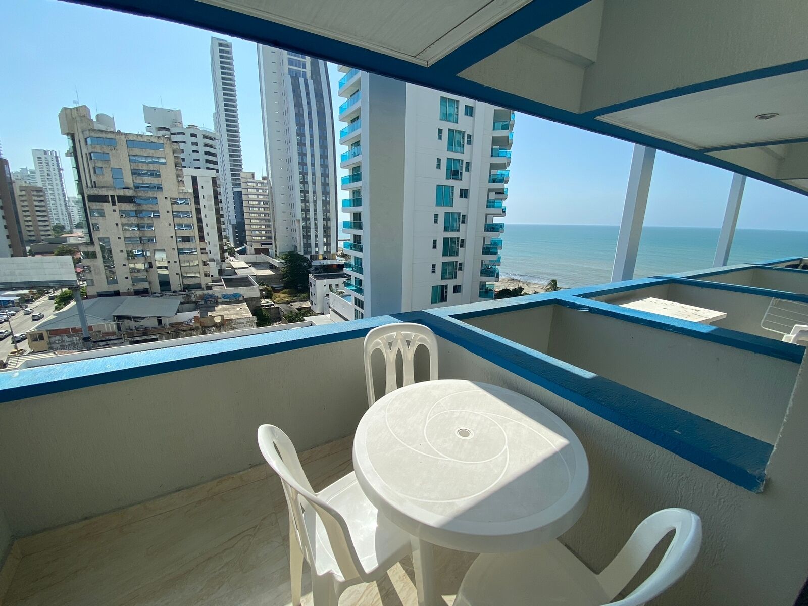 A balcony with a white table and chairs, overlooking a cityscape with tall buildings and a beach.