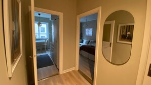Ein Flur, der zu einem Schlafzimmer mit Bett, Schreibtisch und Stuhl führt. An der Wand hängt ein Spiegel und rechts befindet sich eine Tür.