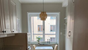 Eine Küche mit weißen Schränken, einem Esstisch mit zwei Stühlen, einem Fenster mit Blick auf Gebäude und einer Pendelleuchte.