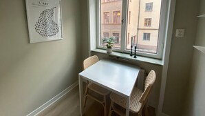 Ein kleiner Raum mit einem weißen Tisch und zwei Korbstühlen, ein Fenster mit Blick auf Gebäude und ein gerahmtes Kunstwerk an der Wand.