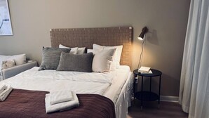 Ein ordentlich bezogenes Bett mit brauner Tagesdecke, weißen Kissen und einem Kopfende mit Backsteinmuster. Ein kleiner Nachttisch mit Lampe und Buch steht daneben.