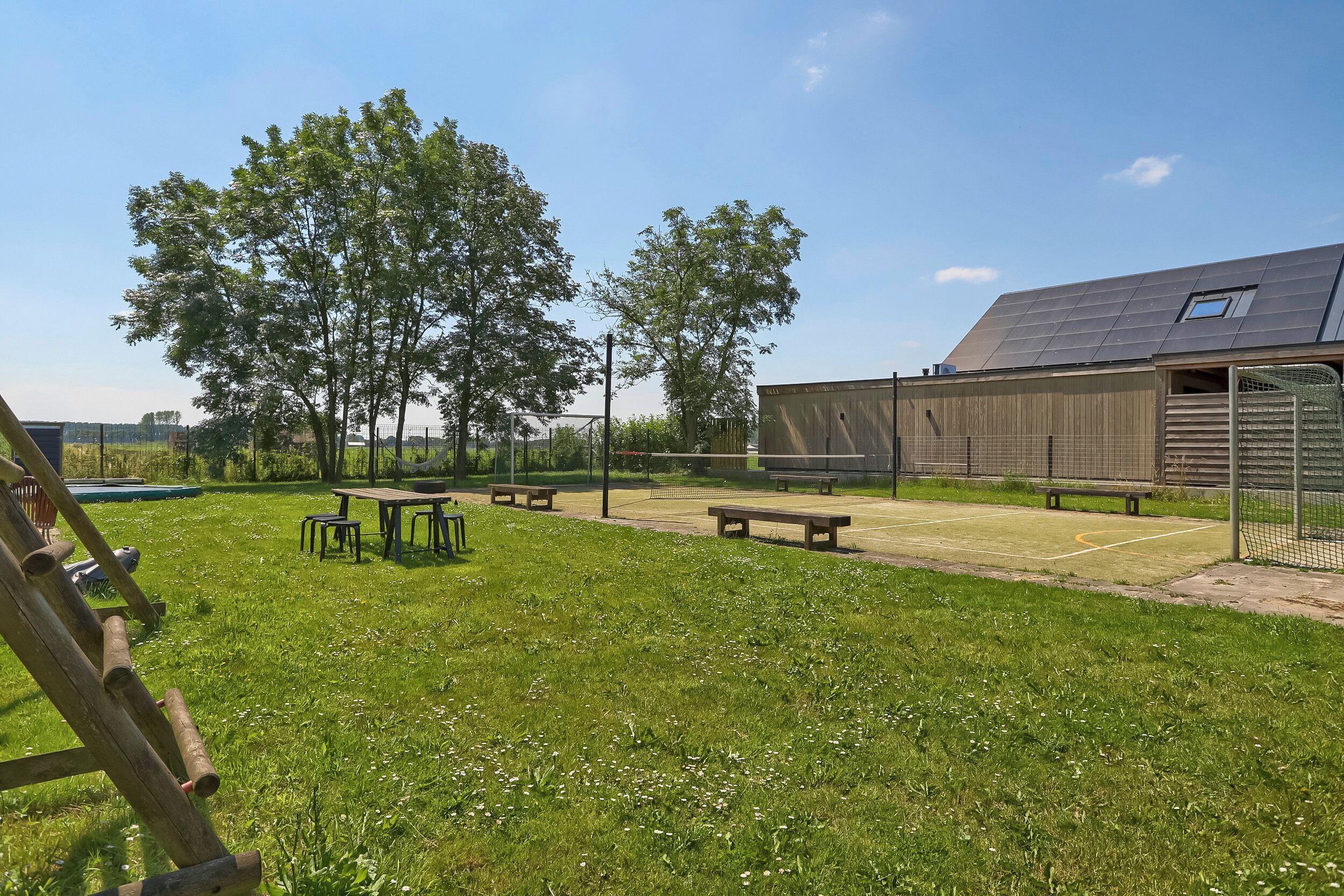 A grassy outdoor area with picnic tables and benches, a wooden building with a sloped roof, and trees in the background.