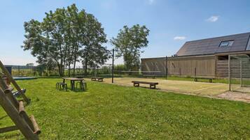 A grassy outdoor area with picnic tables and benches, a wooden building with a sloped roof, and trees in the background.