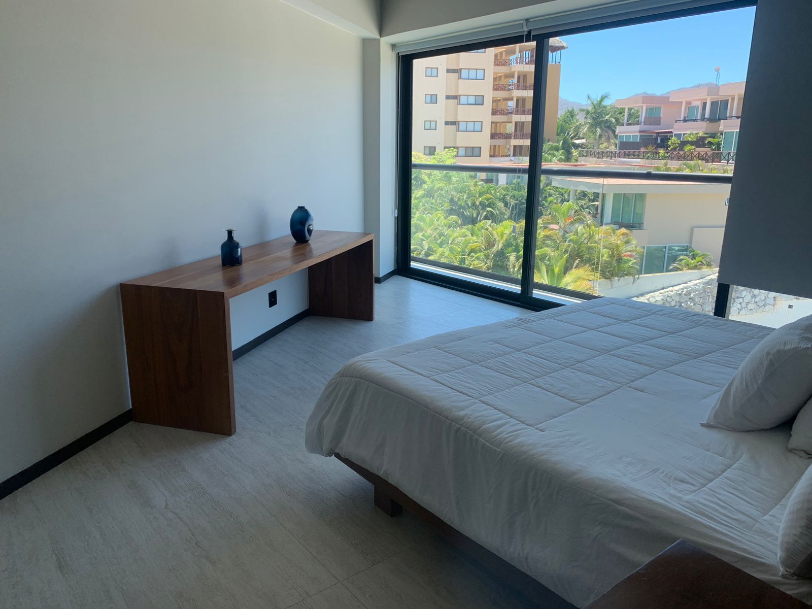 A bedroom with a large bed, a wooden desk, and a window with a view of buildings and greenery.