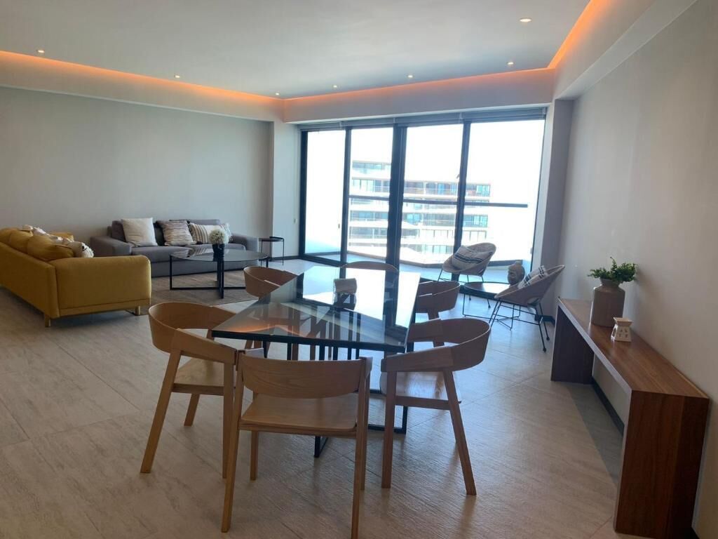 Ein modernes Wohnzimmer mit einem Glastisch, Holzstühlen, einer Couch und einem großen Fenster mit Blick auf Gebäude.