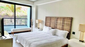Quarto moderno com uma cama grande, duas mesas de cabeceira, um banco e uma janela com vista para áreas verdes.