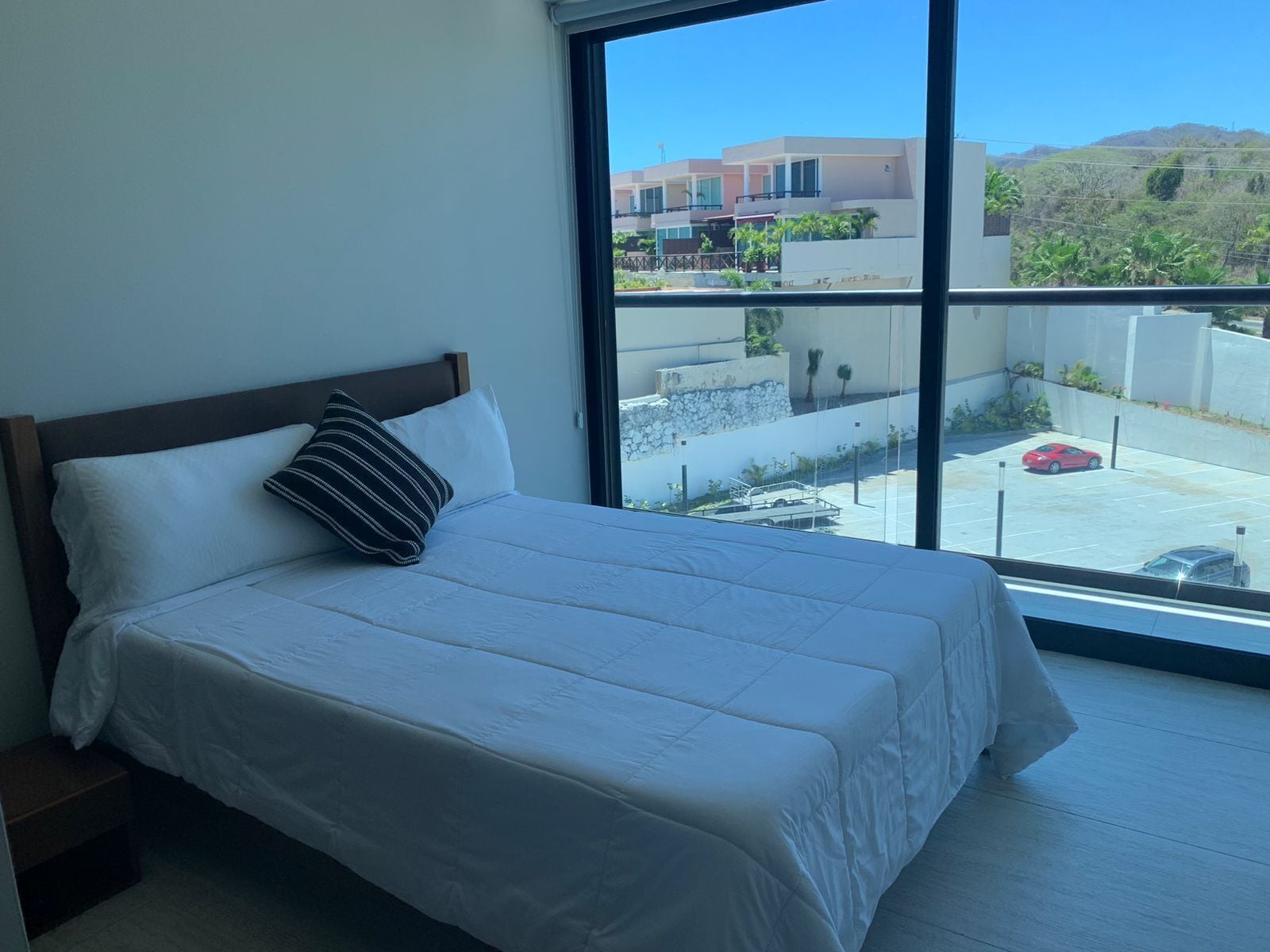 A bedroom with a large window, a bed with white bedding, and a view of a parking area and buildings.