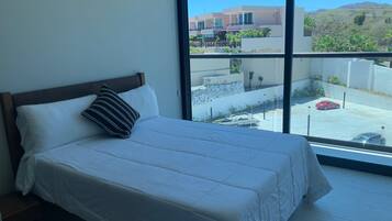 A bedroom with a large window, a bed with white bedding, and a view of a parking area and buildings.