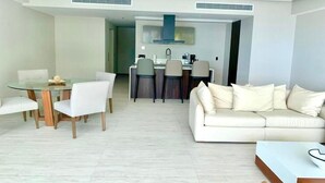 Uma sala de estar moderna com uma mesa de jantar de vidro, sofá branco e uma cozinha ao fundo.