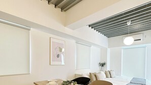 A modern, minimalist room with a wooden dining table set for four, a black chair, a sofa, and a bed with a grey and white duvet. - Arts Dobashi (Hiroshima)