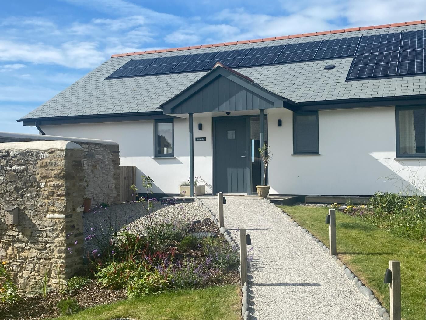 Een modern gebouw met een schuin dak, zonnepanelen, een stenen muur en een grindpad dat naar de ingang leidt.