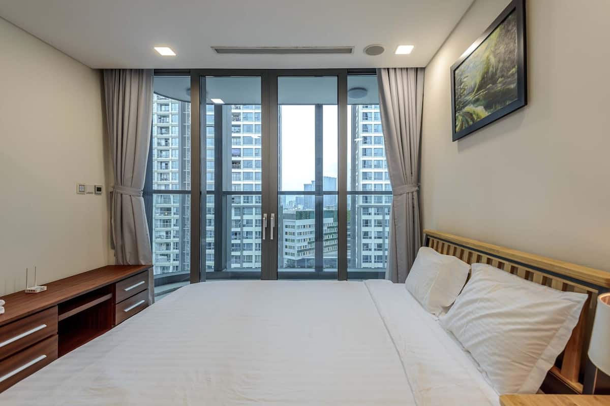 Une chambre d’hôtel avec un grand lit, une commode en bois, un bureau et une vue sur les gratte-ciel, grâce à de grandes fenêtres du sol au plafond.