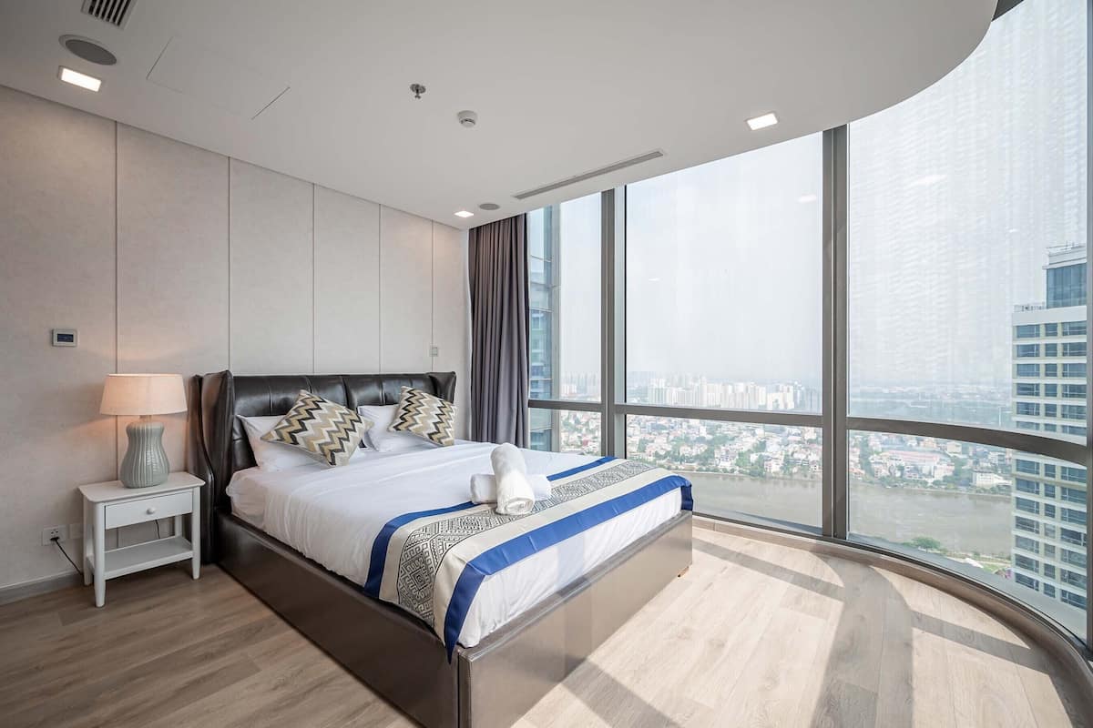 Une chambre moderne avec un grand lit, une table de chevet, une lampe et une vue sur le paysage urbain grâce à de grandes fenêtres.