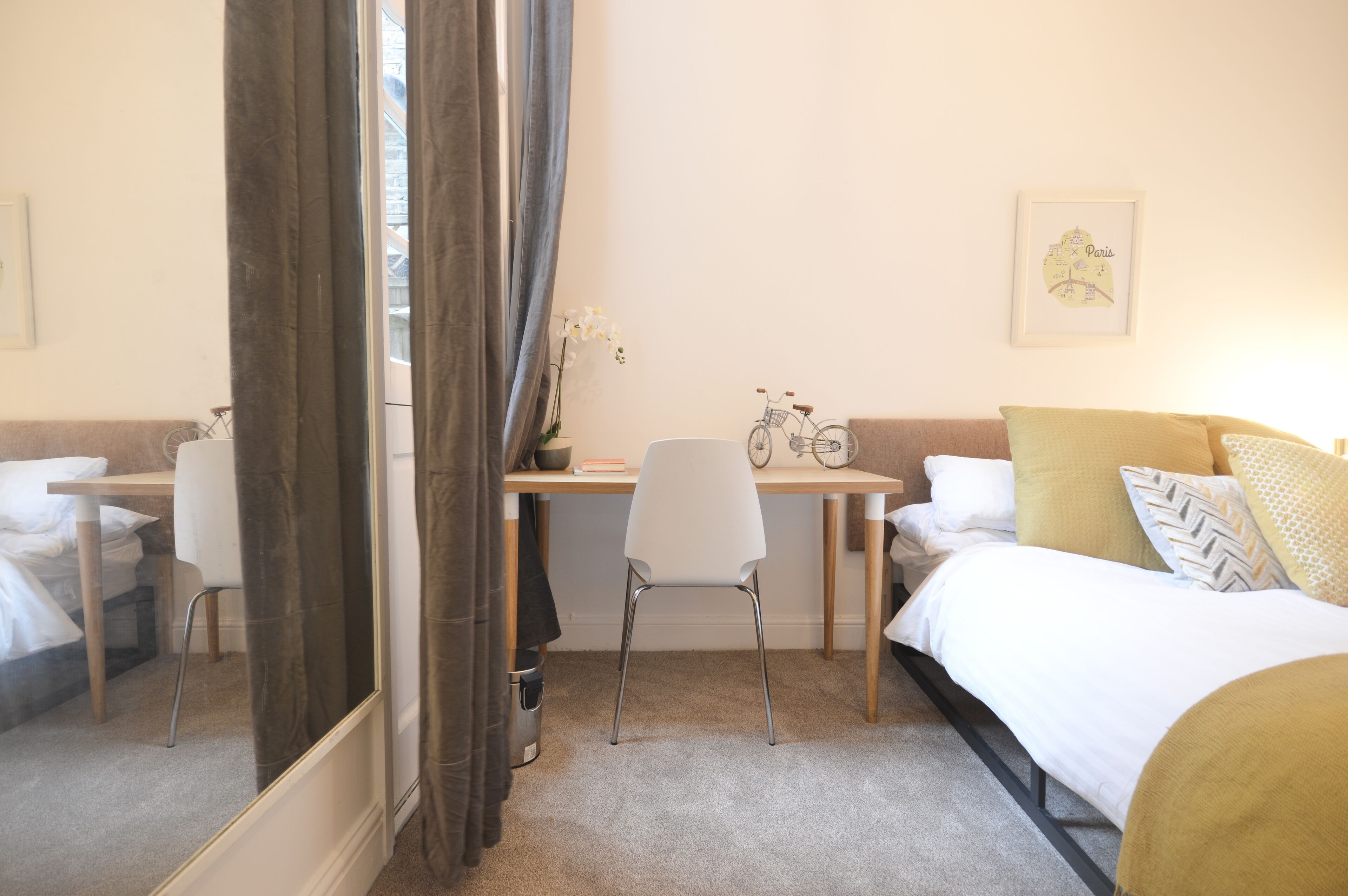 A hotel room with a bed, a desk, a chair, a bicycle, and a framed map of Paris on the wall.