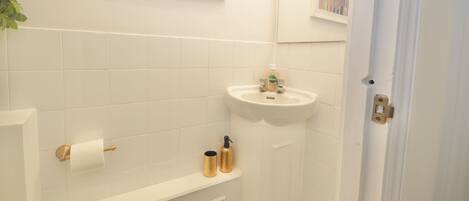 A bathroom with white tiles, a sink, a toilet paper holder, and a shelf with toiletries.