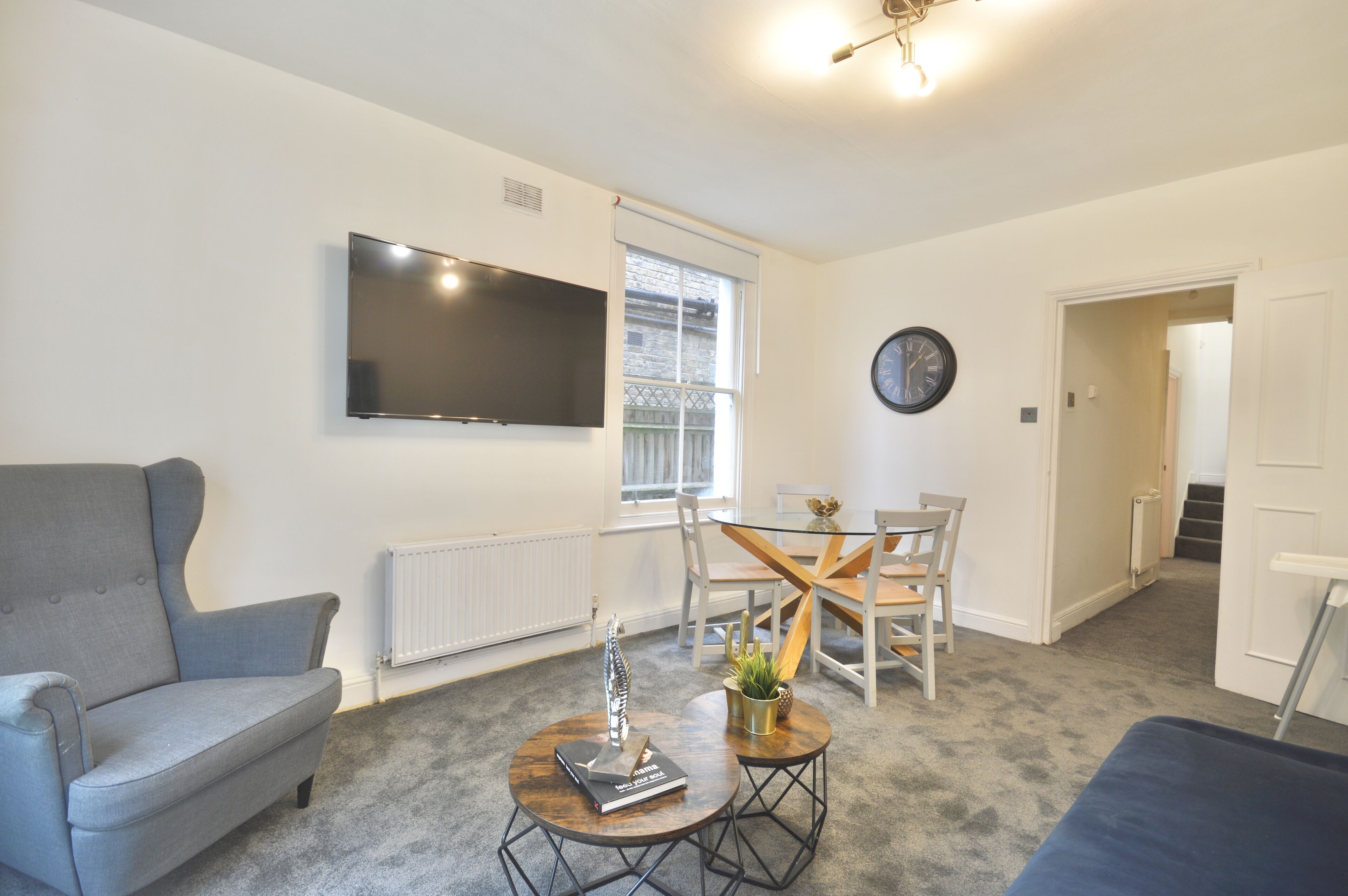 A living room with a grey sofa, a coffee table with a book and a plant, a TV mounted on the wall, and a dining area with a wooden table and chairs.