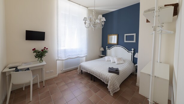 A bedroom with a bed, a desk, a chair, a TV, a window with curtains, a chandelier, and a radiator.