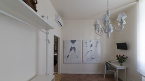 A modern, well-lit room with a white desk, a chair, and a chandelier. There are two abstract paintings on the wall and a television mounted on the wall.