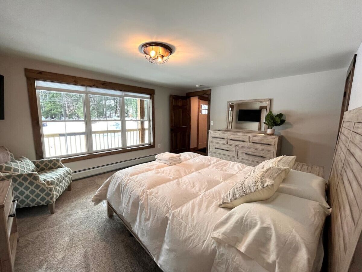 A bedroom with a large bed, a dresser, a mirror, and a window with a view of trees.
