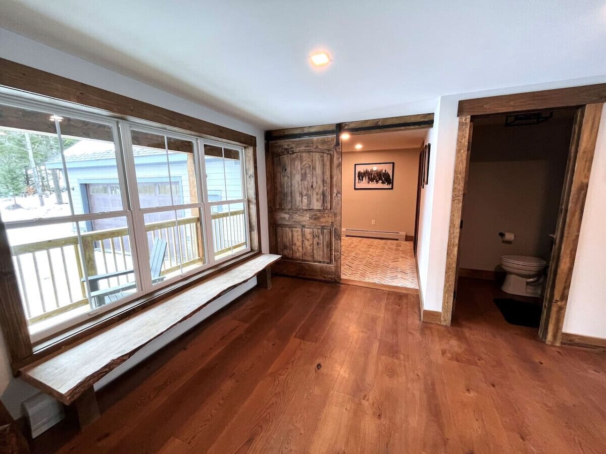 Una habitación con suelo de madera, un banco junto a un ventanal grande y una puerta que da al baño.