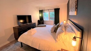 Un dormitorio con cama, televisor, cómoda y ventana con cortinas.
