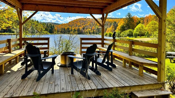 Un pontile di legno con due poltrone Adirondack e un tavolino, con vista su un lago e il fogliame autunnale.