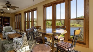 Zona pranzo con tavolo rotondo in legno e sei sedie, ampie finestre vista lago e ventilatori a soffitto.