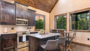 A kitchen with dark wooden cabinets, stainless steel appliances, a marble island, and large windows offering a view of trees. - Haus of Pine Cabin | Modern Woodland Cabin (Hochatown)