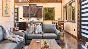 A spacious living room with a dark leather sofa and a coffee table, a kitchen with wooden cabinets and a microwave, and large windows offering a view of trees. - Haus of Pine Cabin | Modern Woodland Cabin (Hochatown)