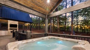 A hot tub area with a stone fireplace, wooden deck, and a view of the forest. - Haus of Pine Cabin | Modern Woodland Cabin (Hochatown)