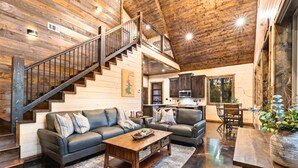 A spacious living room with a staircase, leather sofas, a wooden coffee table, and a kitchen area. - Haus of Pine Cabin | Modern Woodland Cabin (Hochatown)