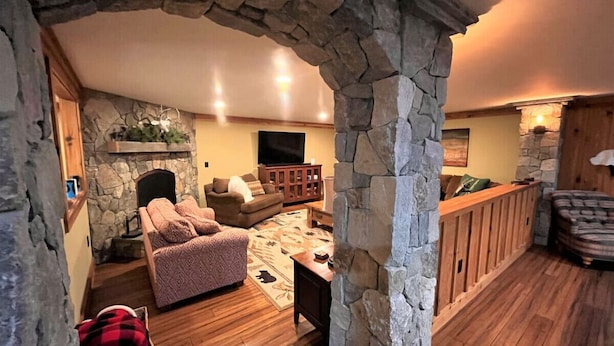 A cozy living room with a stone fireplace, wooden flooring, and a stone archway. - New! Min from Mt & Town. Hot tub, Stream + privacy (Wilmington)