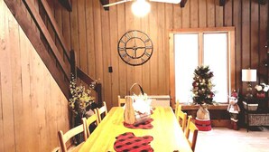 Een houten eetkamer met een lange tafel, stoelen, een plafondventilator, een wandklok en een kerstboom.