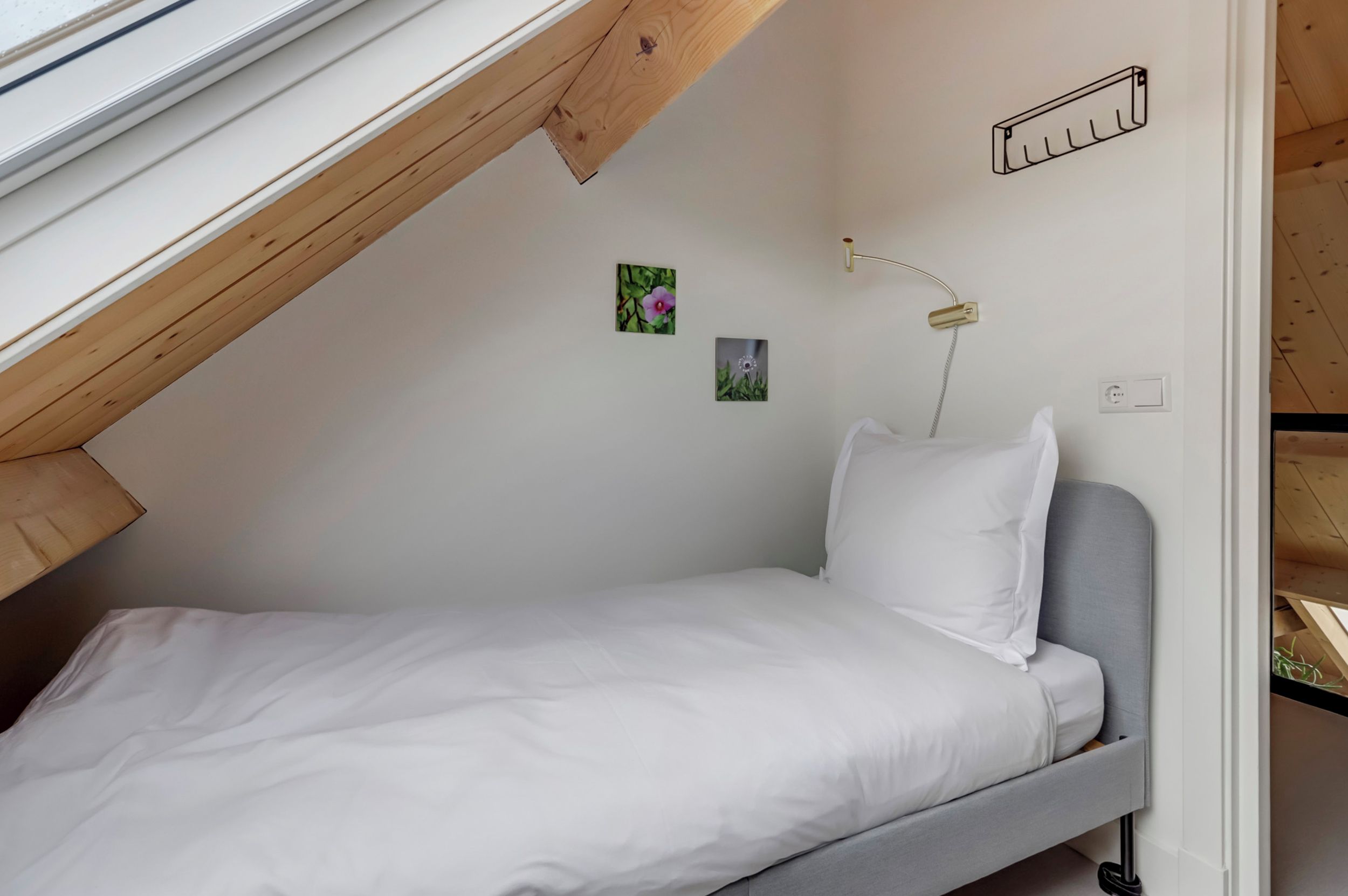 Ein kleines Zimmer mit einem Einzelbett, geneigter Decke, Nachttisch, Wandlampe und zwei kleinen Wandbildern.