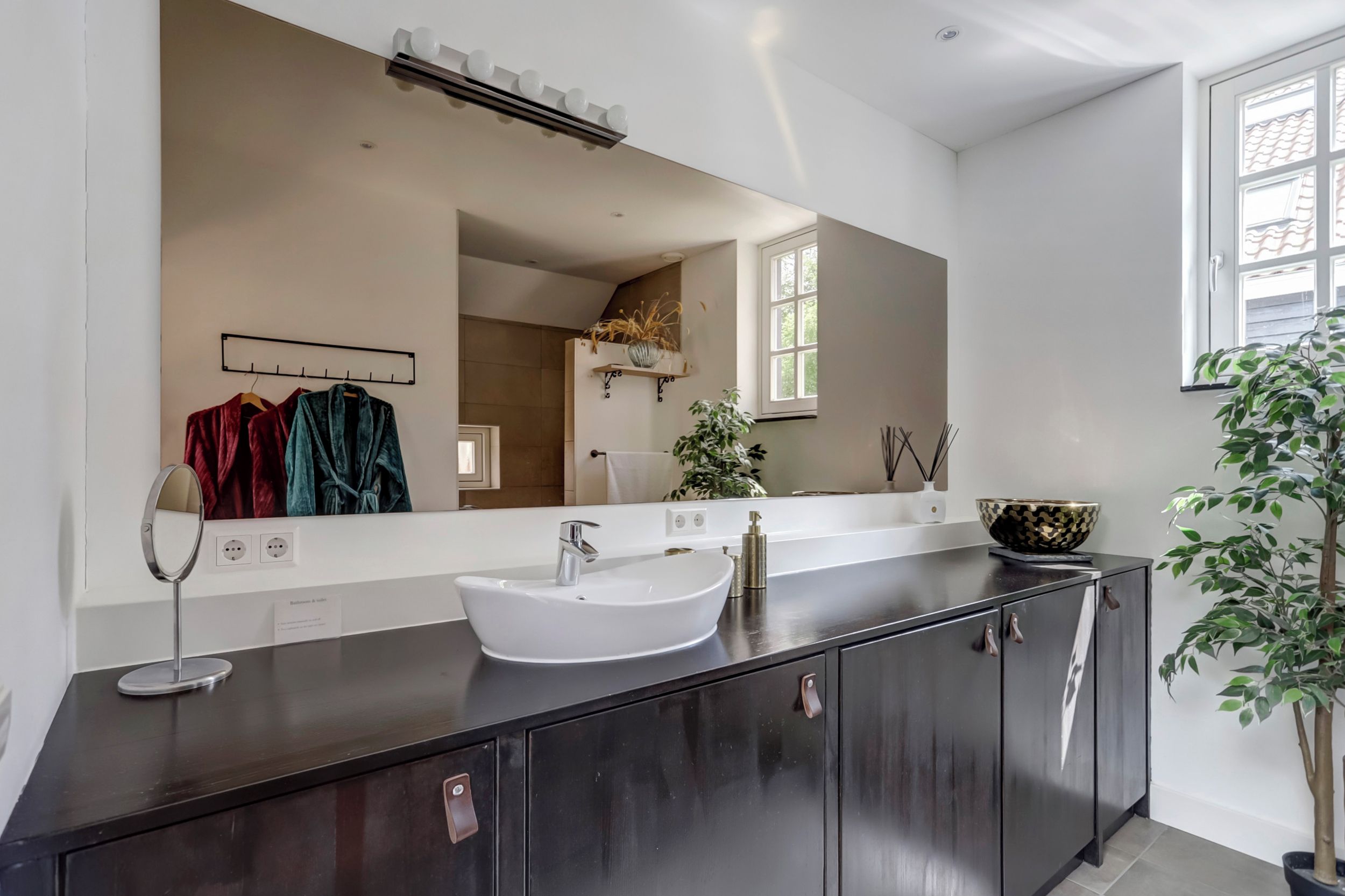 Un baño moderno con un espejo grande, un lavamanos blanco y un mueble oscuro.