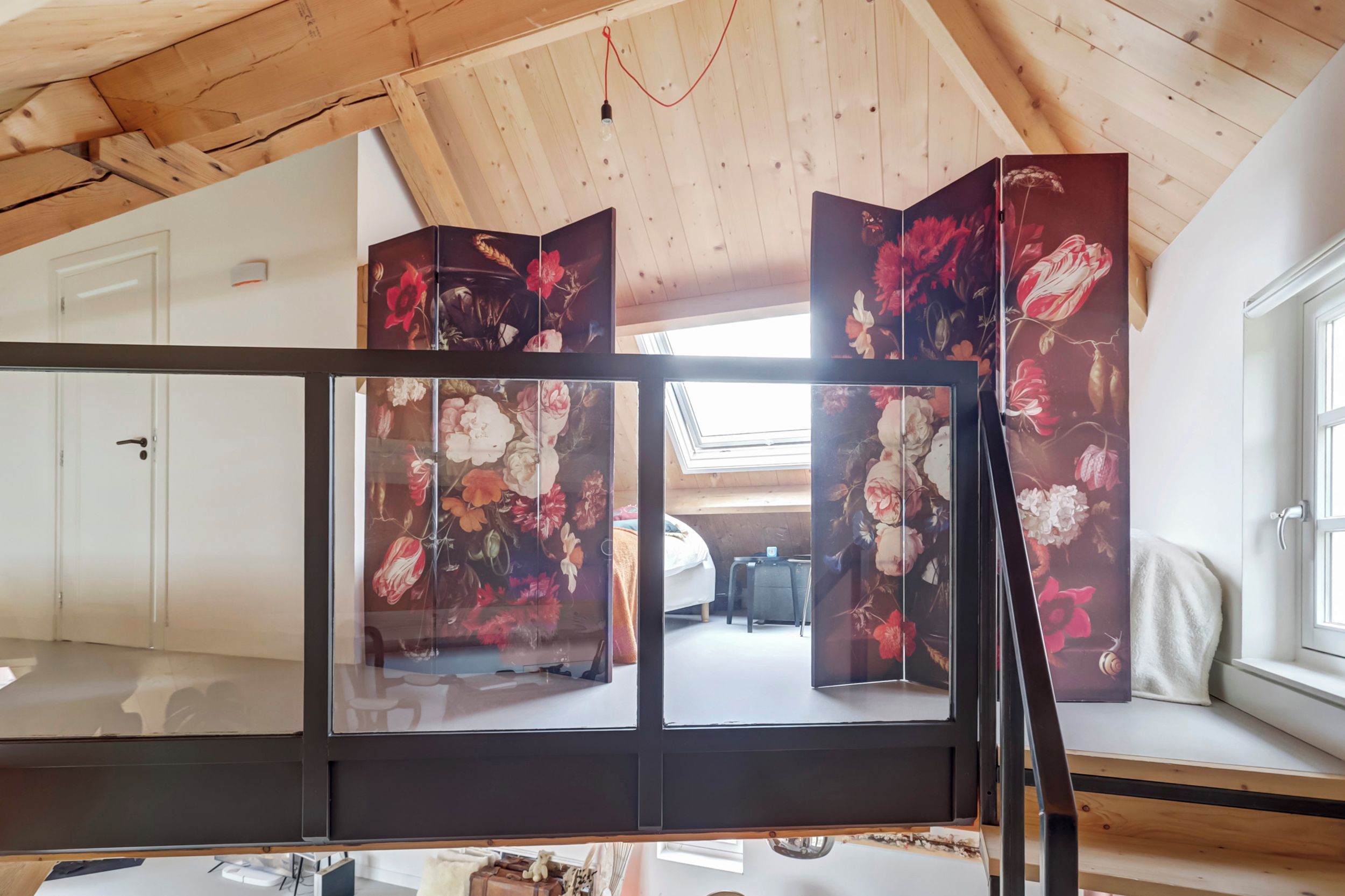 Ein Zimmer mit schrägen Wänden, einem Bett, einem Schreibtisch und einem großen Fenster.