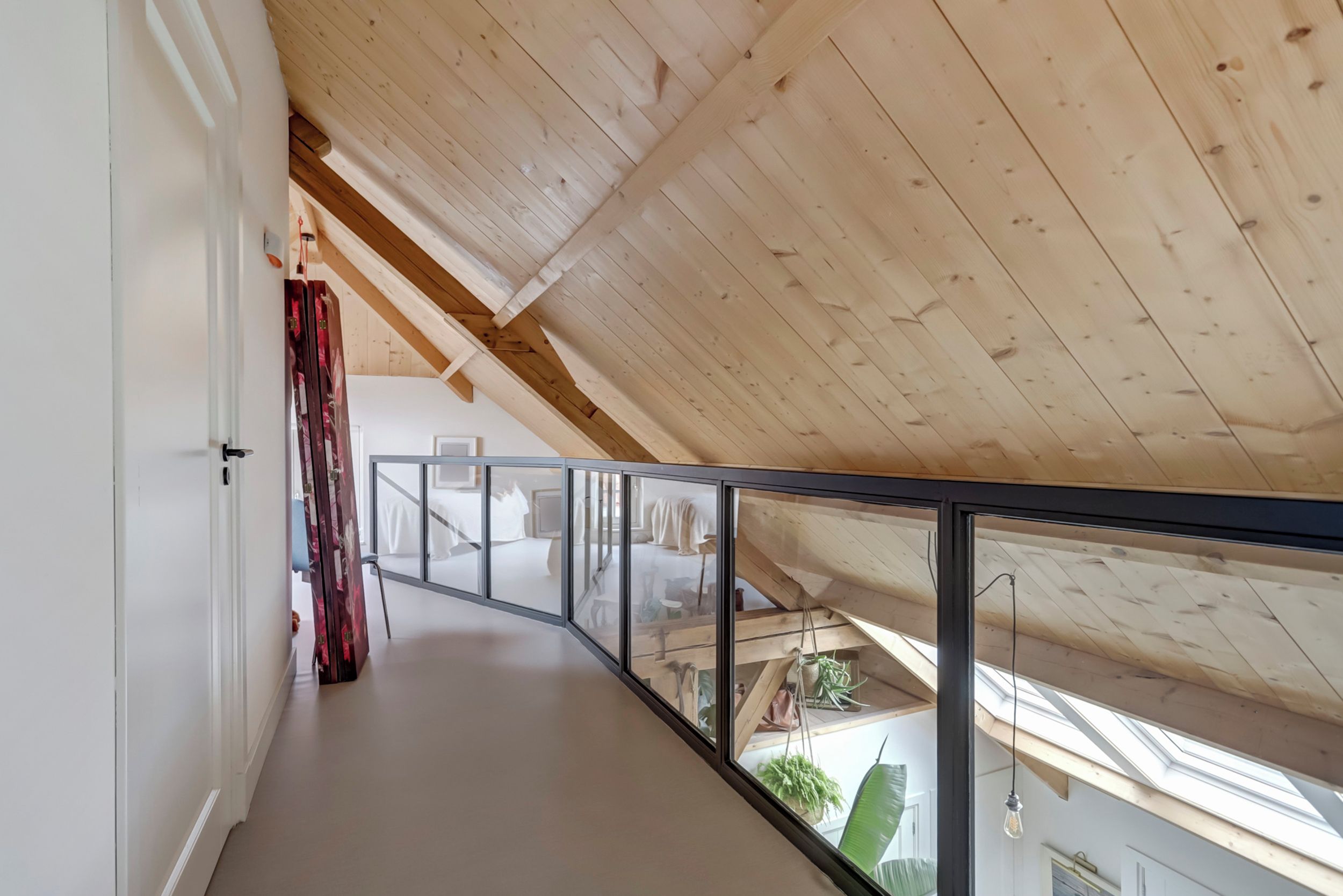 Un couloir doté d’un plafond en bois et d’une rambarde en verre, qui mène à une chambre à coucher avec un lit et une fenêtre.