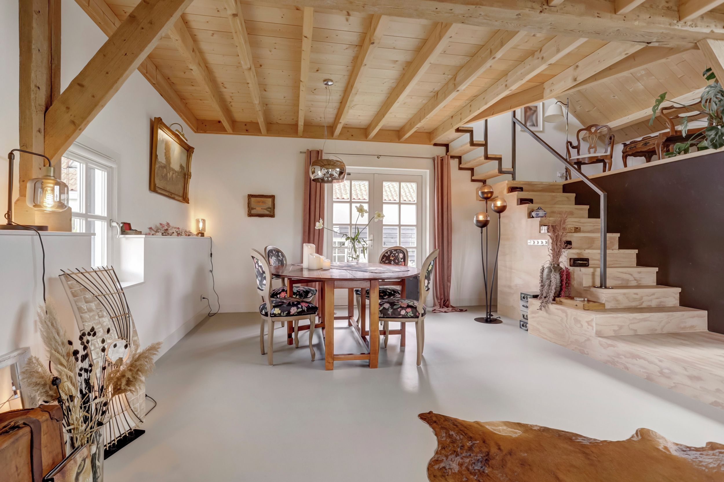 A spacious room with a wooden ceiling, a dining table with chairs, a staircase, and a window with curtains.