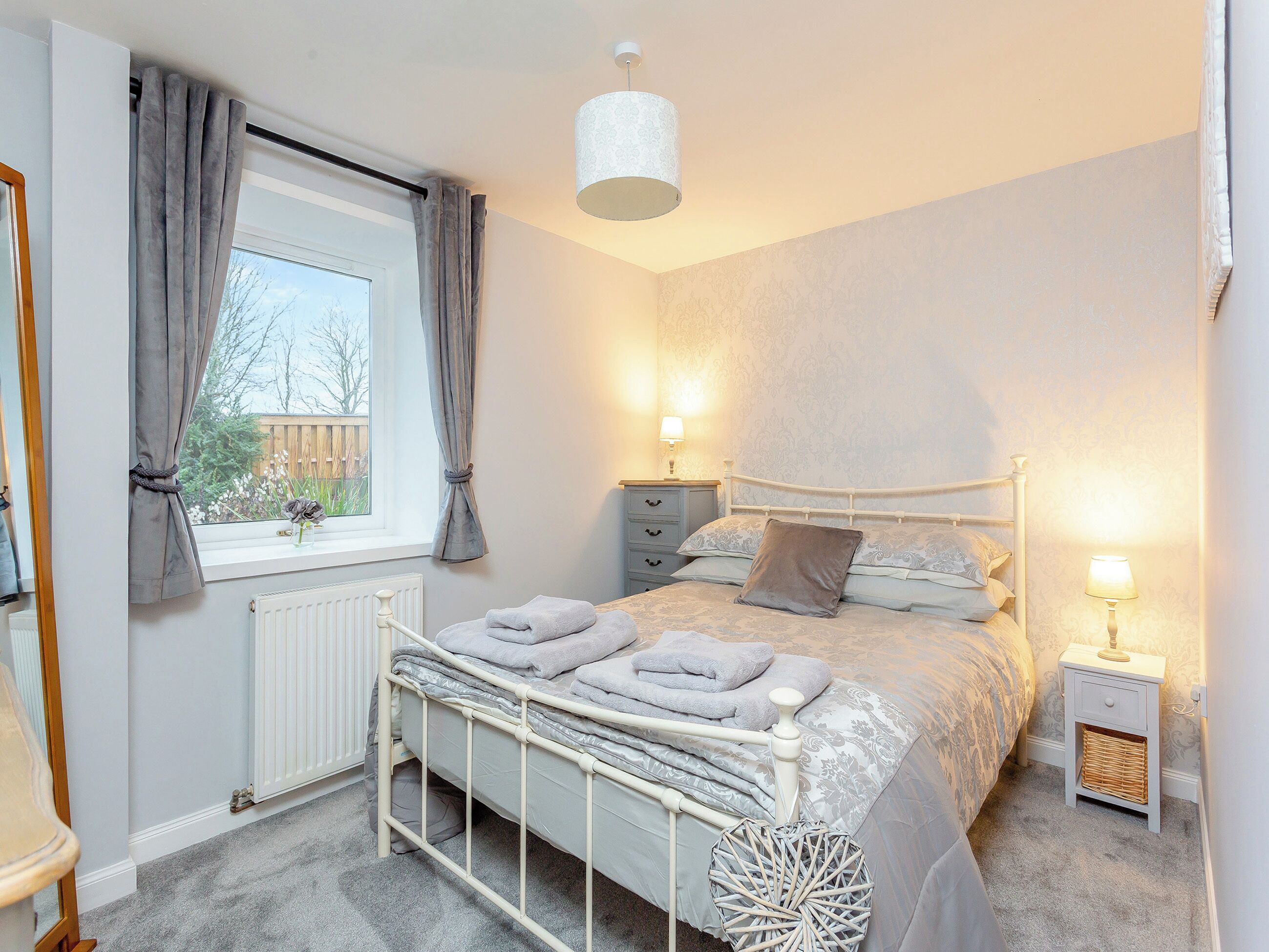 A bedroom with a bed, bedside tables, a window with curtains, a radiator, and a hanging light fixture.