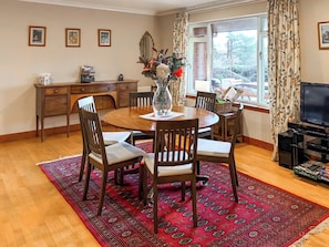 A dining area with a round wooden table, four chairs, a red patterned rug, a television, and a window with curtains. - 3 bedroom accommodation in Alyth (Alyth)