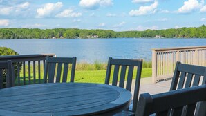 Una terraza de madera con una mesa redonda y sillas, con vista a un lago bajo un cielo despejado y una orilla distante.